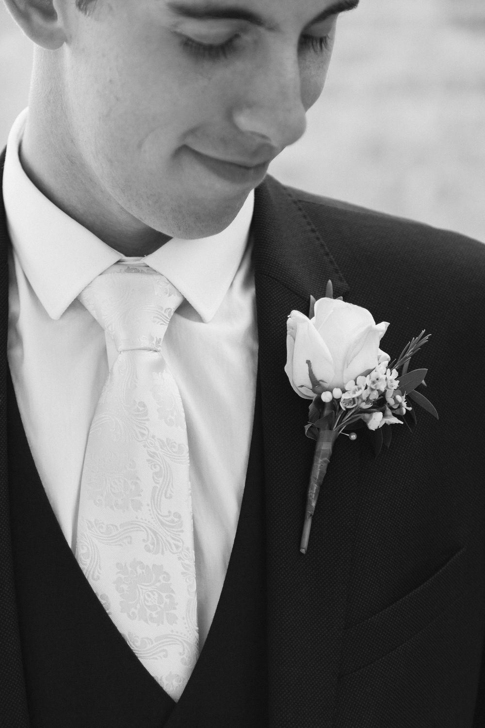 Close-up of groom wearing classic black suit with white tie and rose boutonniere in elegant black and white photo.