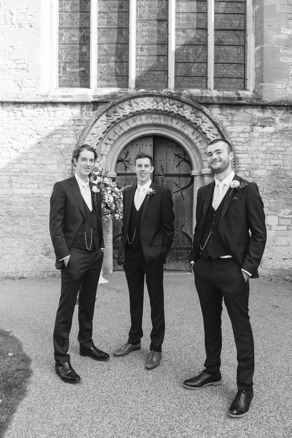 Three groomsmen in matching dark suits pose in front of ornate church doorway.