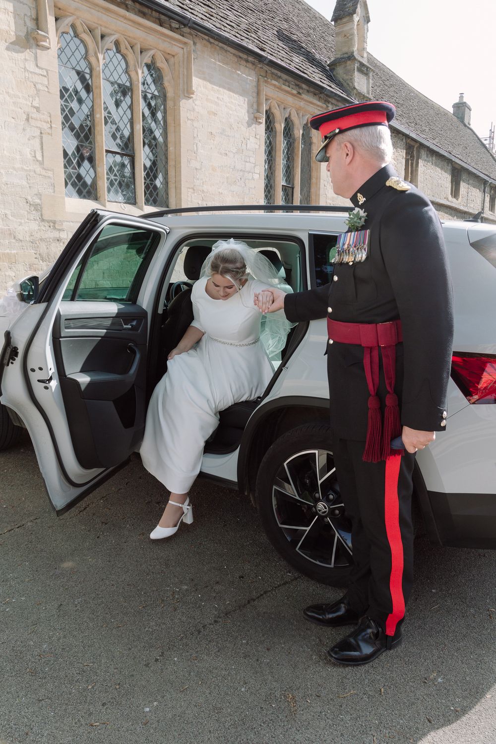 A person in a white dress exits a car while being assisted by a uniformed officer in front of a stone church.