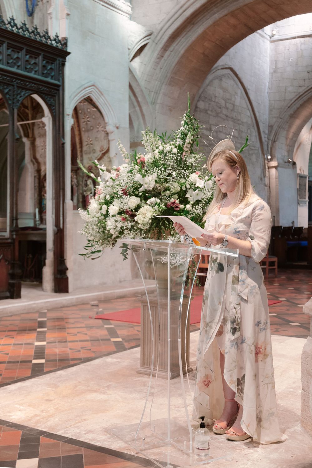 Someone stands at a clear podium with elegant floral arrangements in a historic church with vaulted arches and tiled floors.