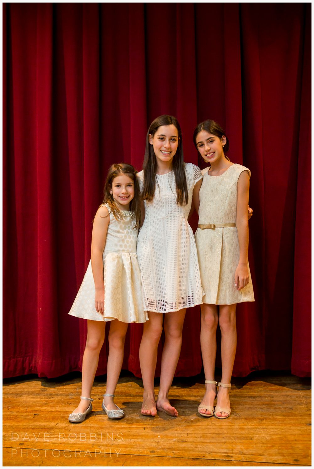 Three pairs of feet and white dresses stand against a red curtain backdrop on a wooden stage.