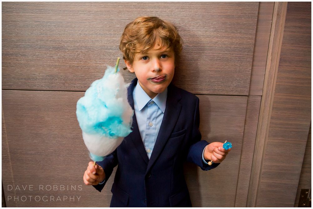 Young child in a navy blue suit and tie makes silly faces while holding cotton candy at a formal event.