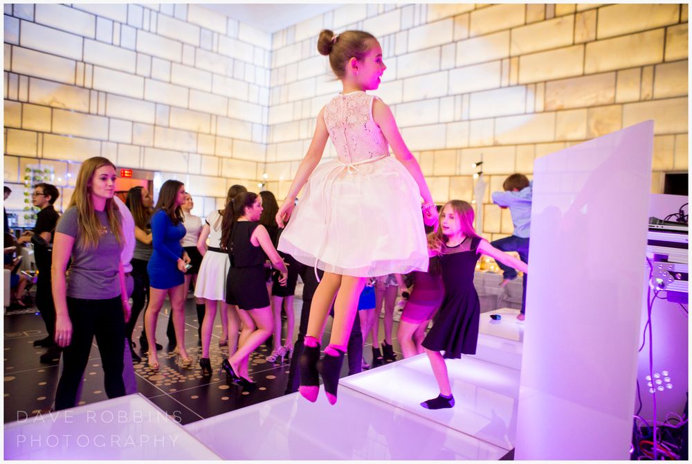 People dancing and celebrating on a white tiled floor with purple ambient lighting at an event venue.