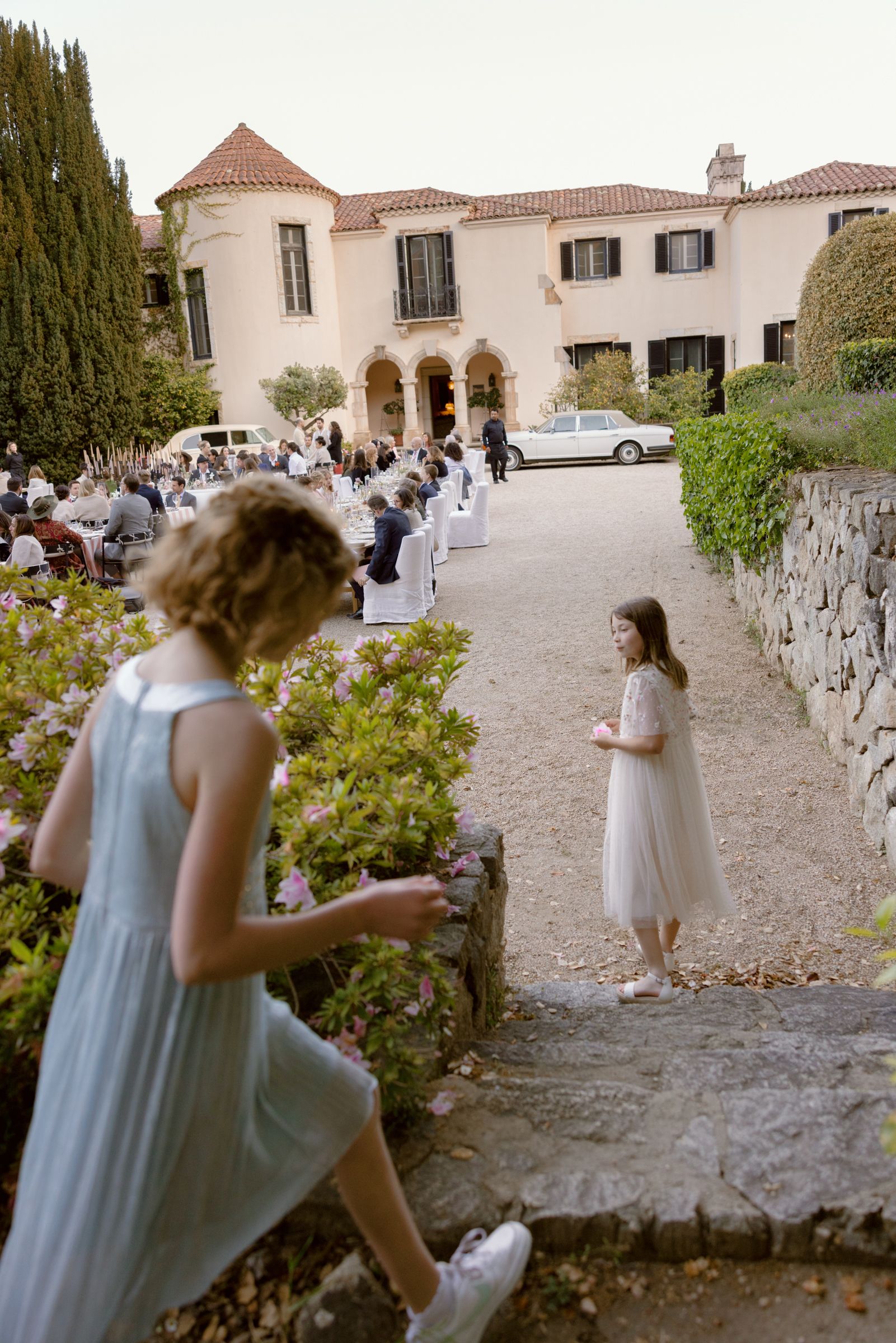 Outdoor wedding celebration at Mediterranean-style villa with white chairs and guests on gravel driveway.