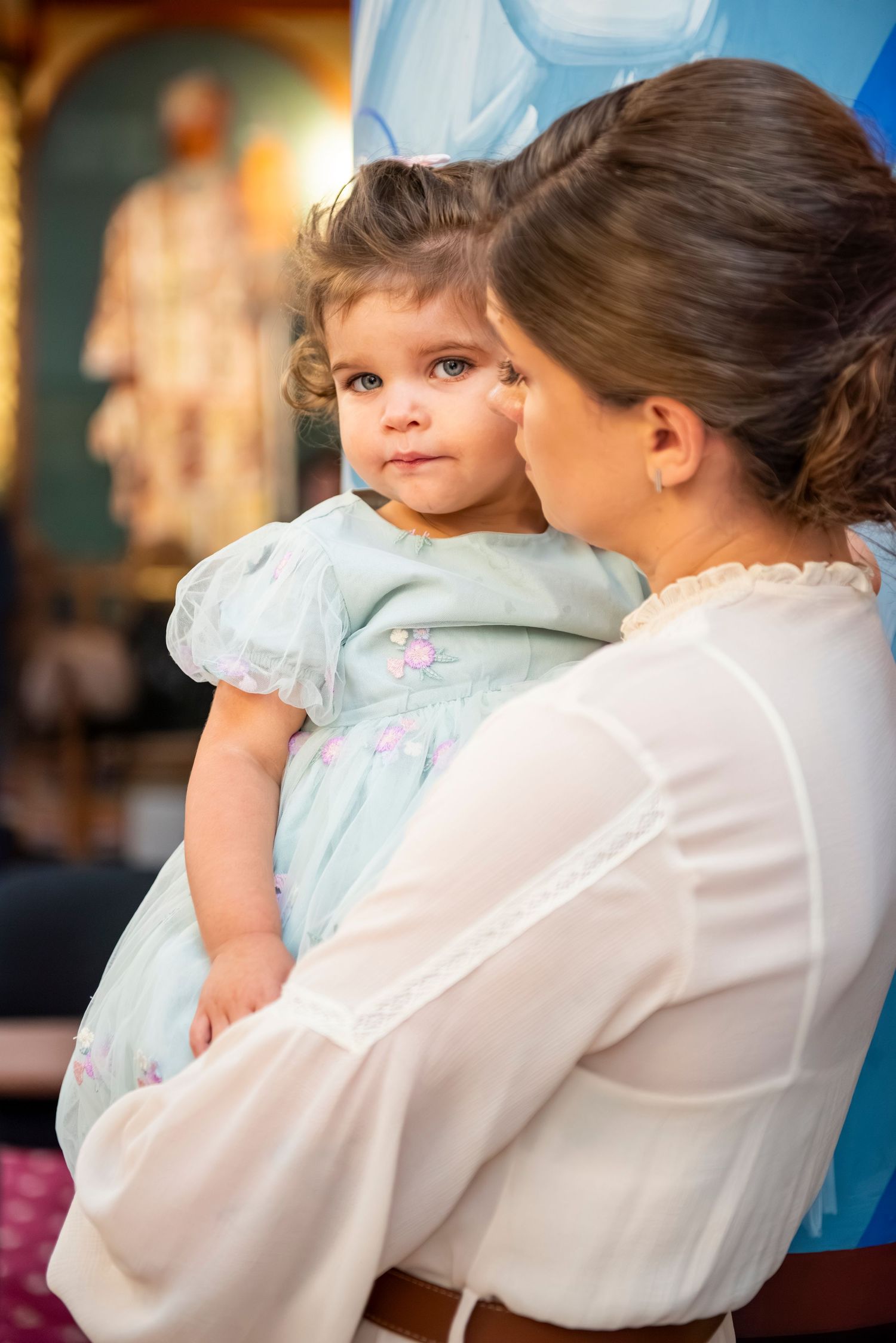 Parent in white blouse holds small child wearing light blue dress.