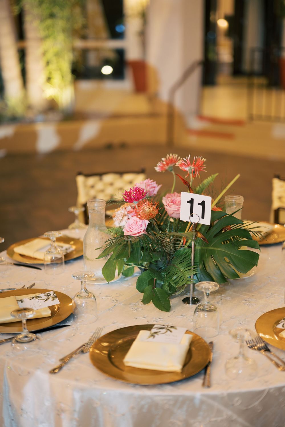 Elegant table settings with gold charger plates and tropical floral centerpieces featuring monstera leaves and pink flowers.