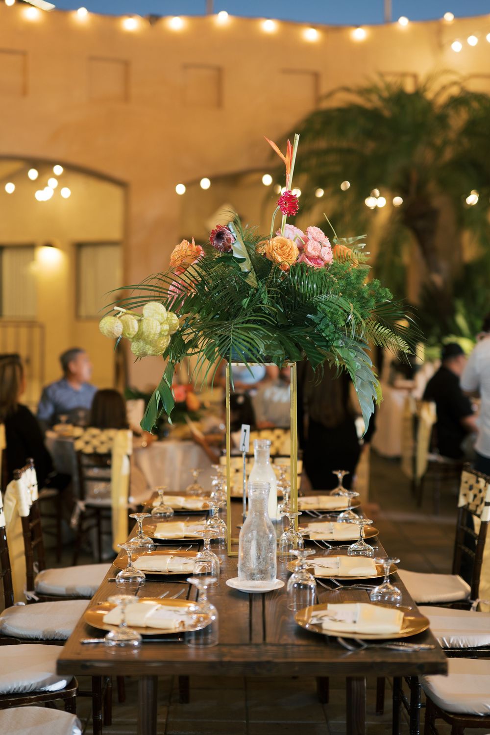 Tall tropical centerpiece adorns wedding reception table under string lights in an elegant indoor venue.