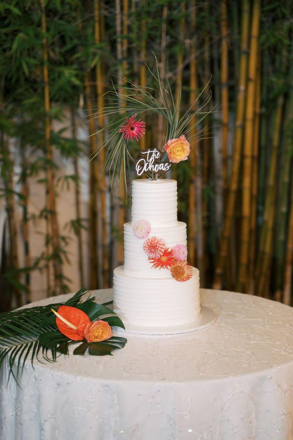 Three-tiered white wedding cake decorated with tropical flowers and monstera leaves displayed against bamboo backdrop.