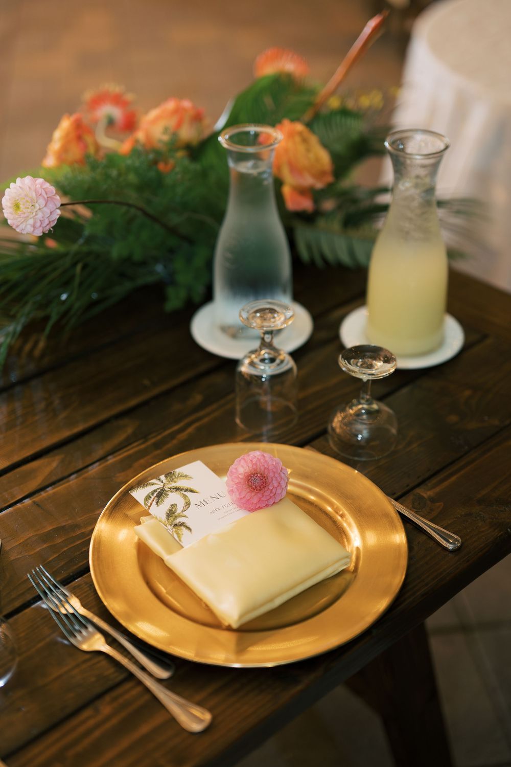 Elegant table setting with gold charger plate, pink flower, and glass bottles of drinks on rustic wooden table.