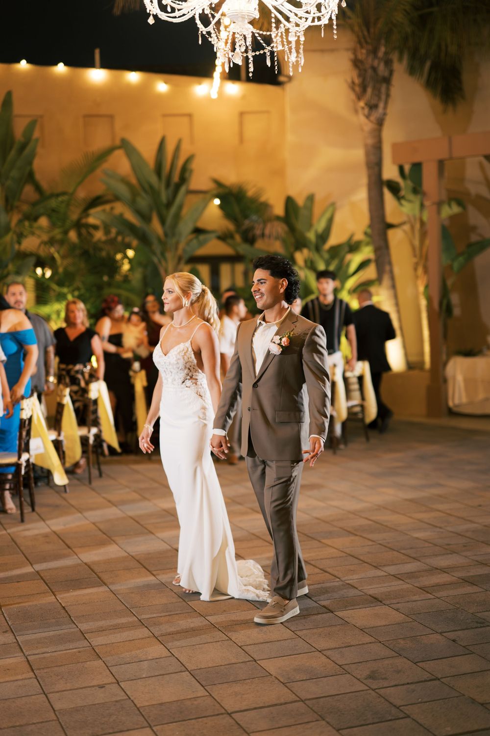 Elegant outdoor wedding reception with couple walking under a crystal chandelier surrounded by tropical plants.
