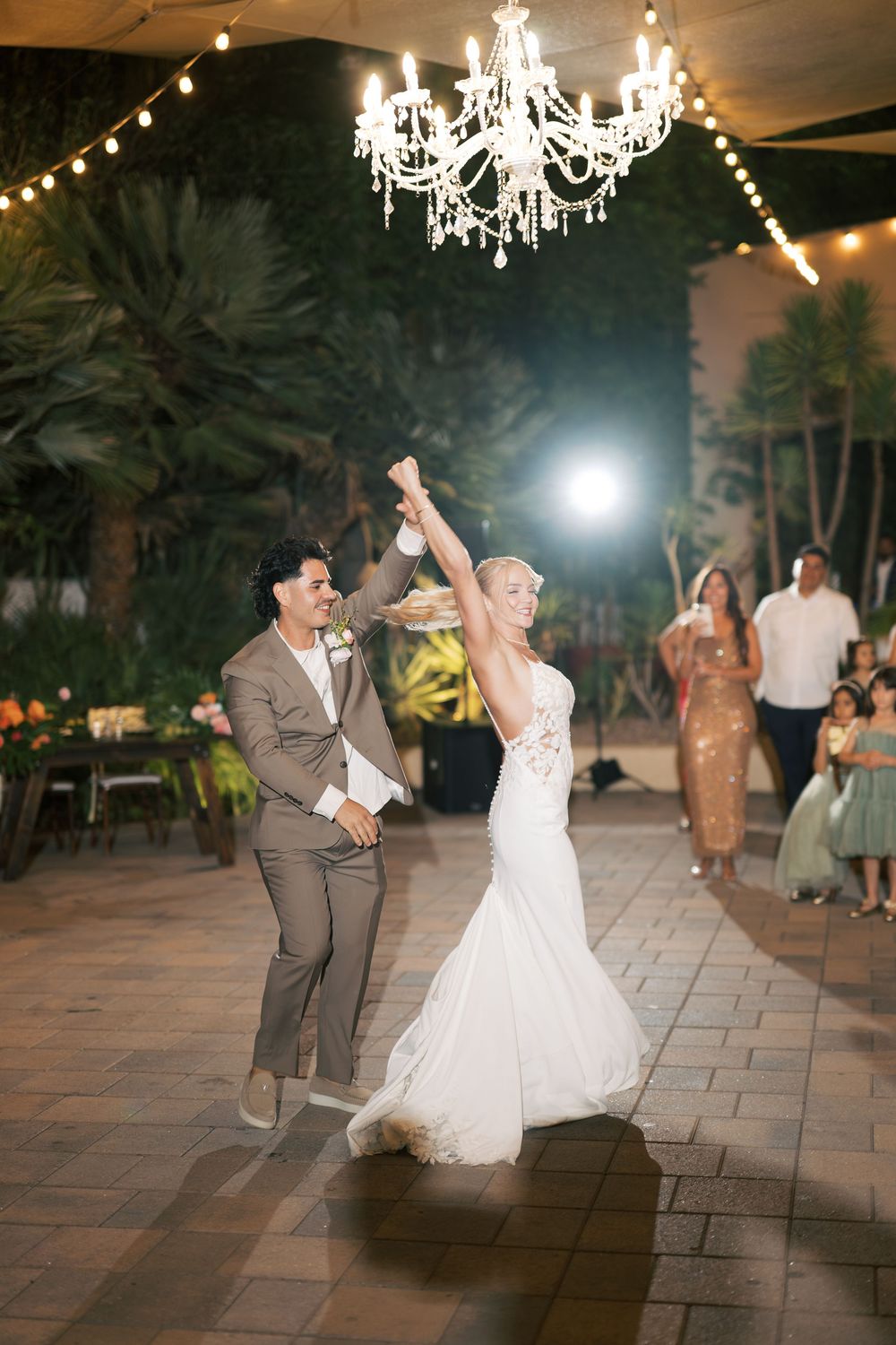 Wedding couple dances under crystal chandeliers and string lights on an outdoor patio at night.