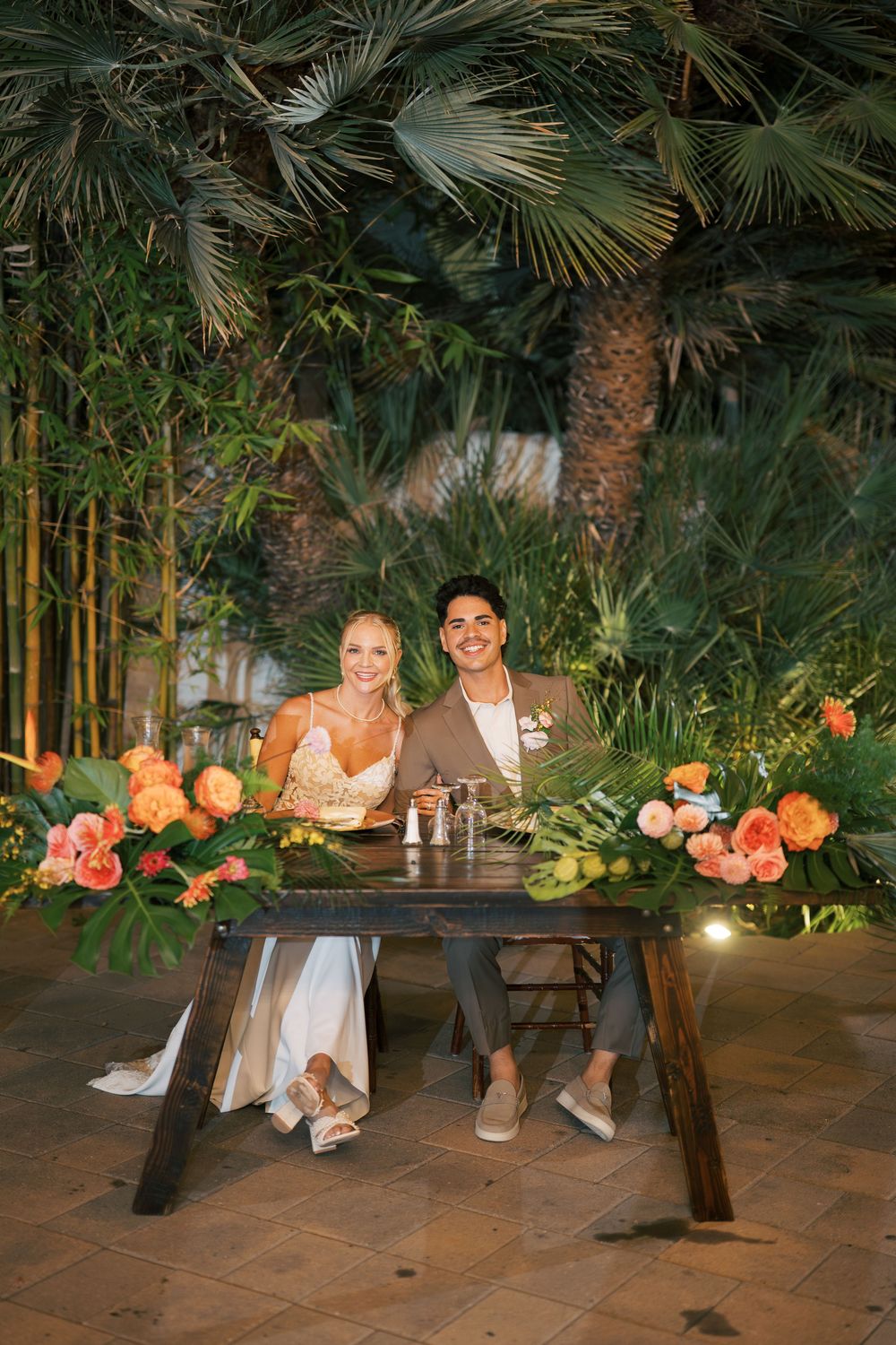 Wedding couple seated at rustic sweetheart table decorated with tropical flowers and palm fronds.