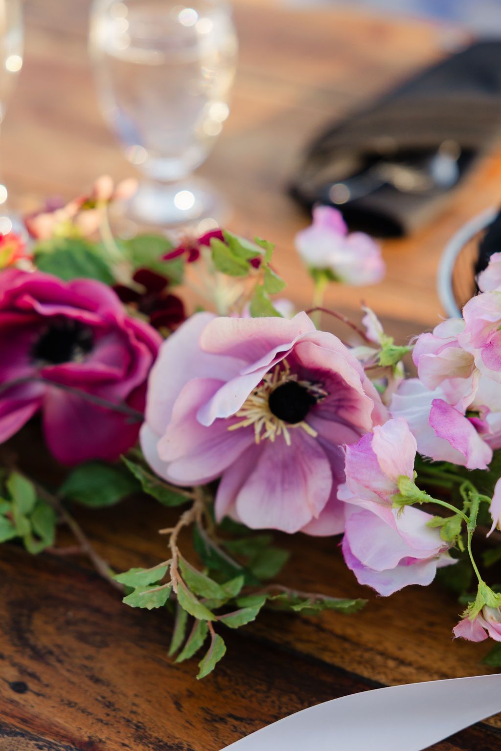Delicate pink and purple anemone flowers arranged on a rustic wooden surface with soft bokeh lighting in the background.