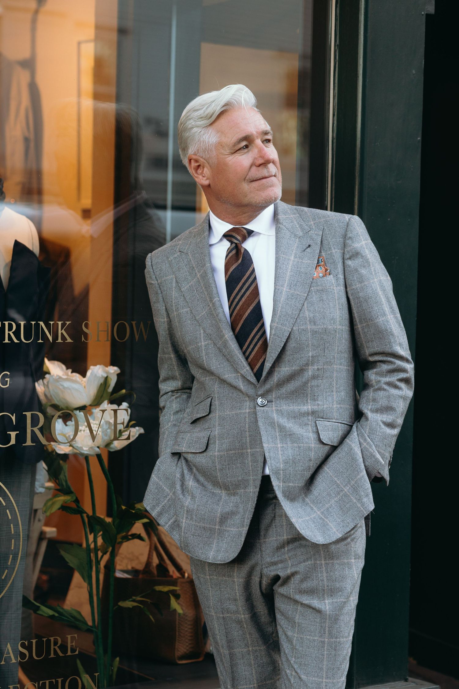 Distinguished figure in grey plaid suit posing against storefront window.
