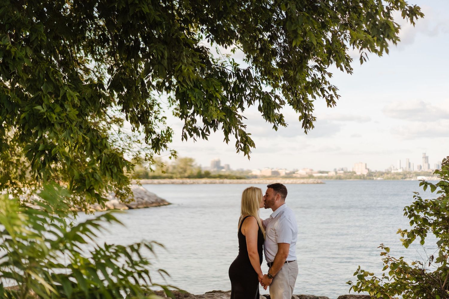 A romantic couple embraces by the water's edge under tree branches at sunset, creating a serene outdoor scene.