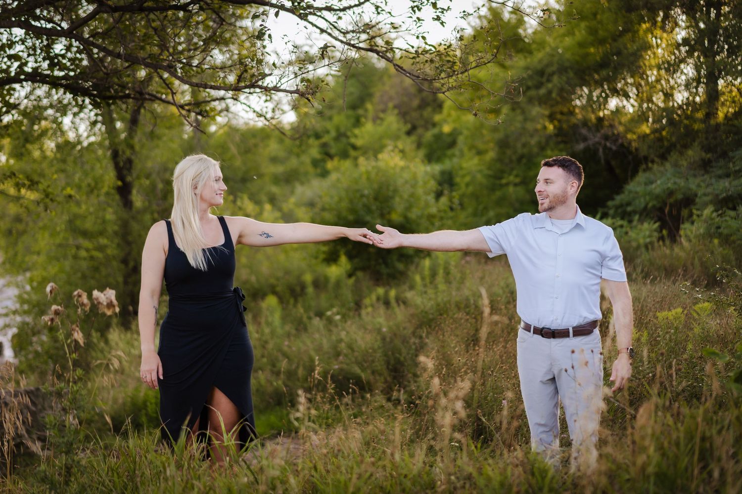 A couple walks together among tall grass and trees in a scenic outdoor setting during golden hour lighting.