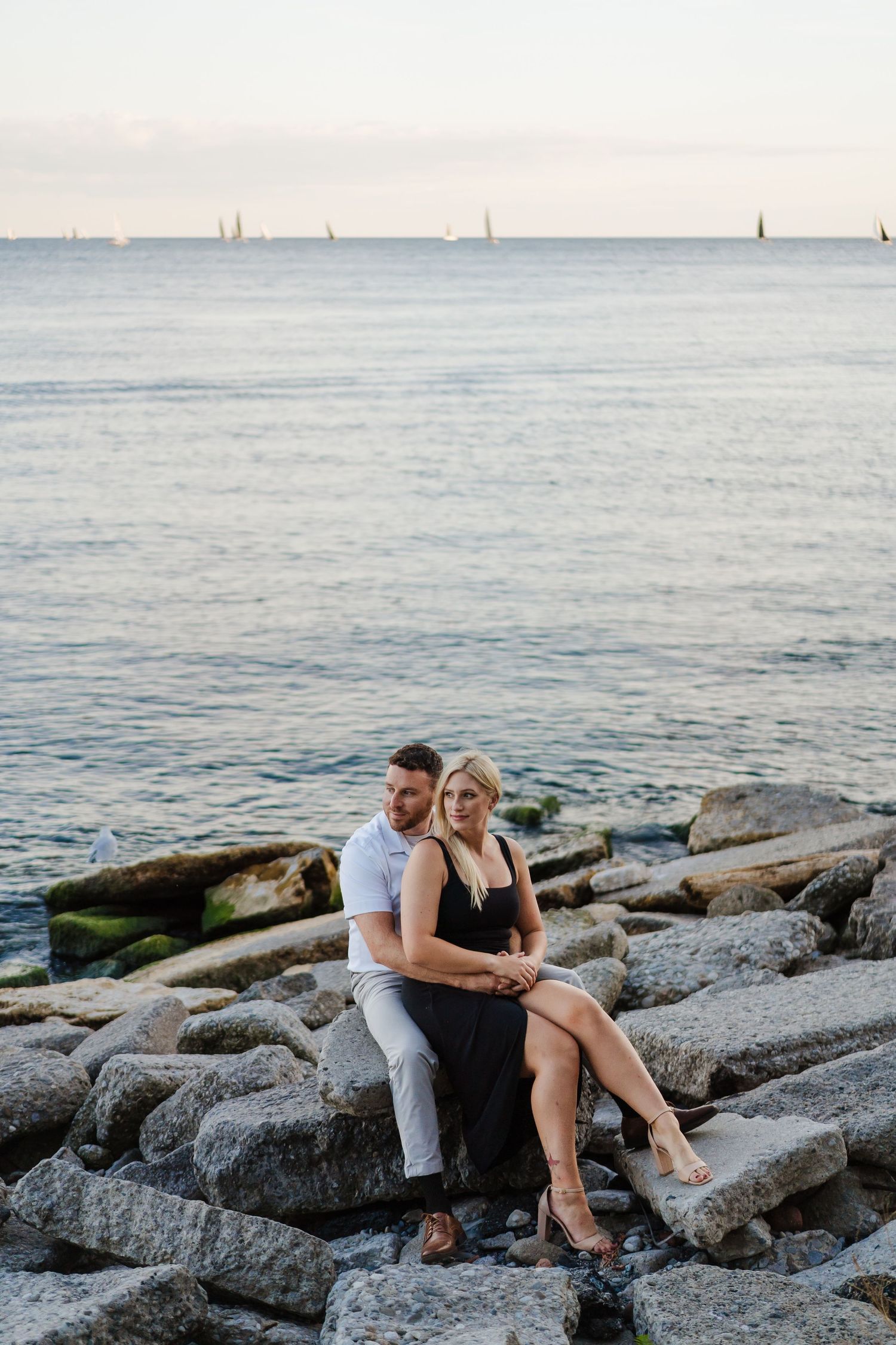 Couple sitting together on rocky shoreline with ocean waves at sunset.