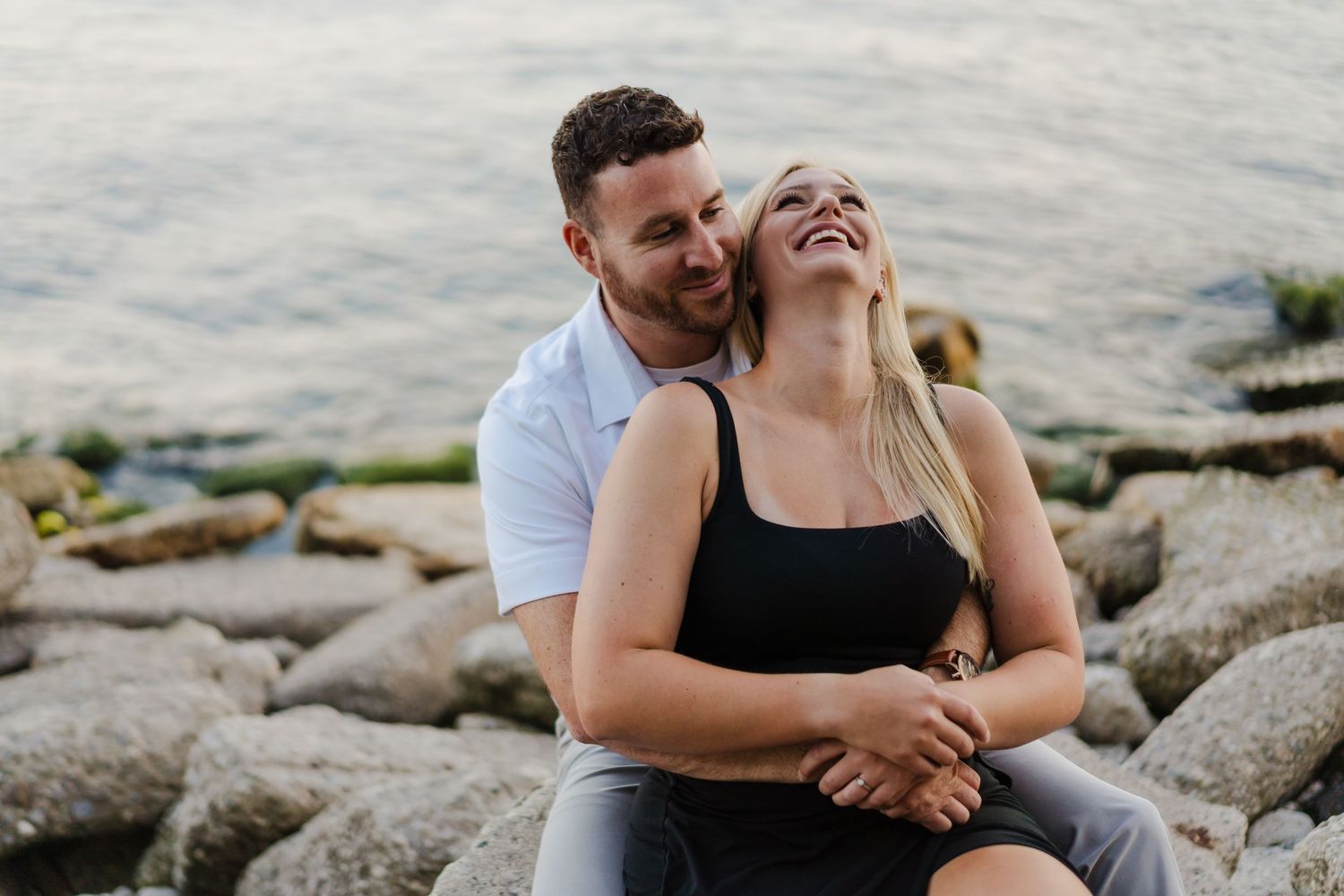 Happy couple laughing together while sitting on rocky beach shore during romantic sunset moment.