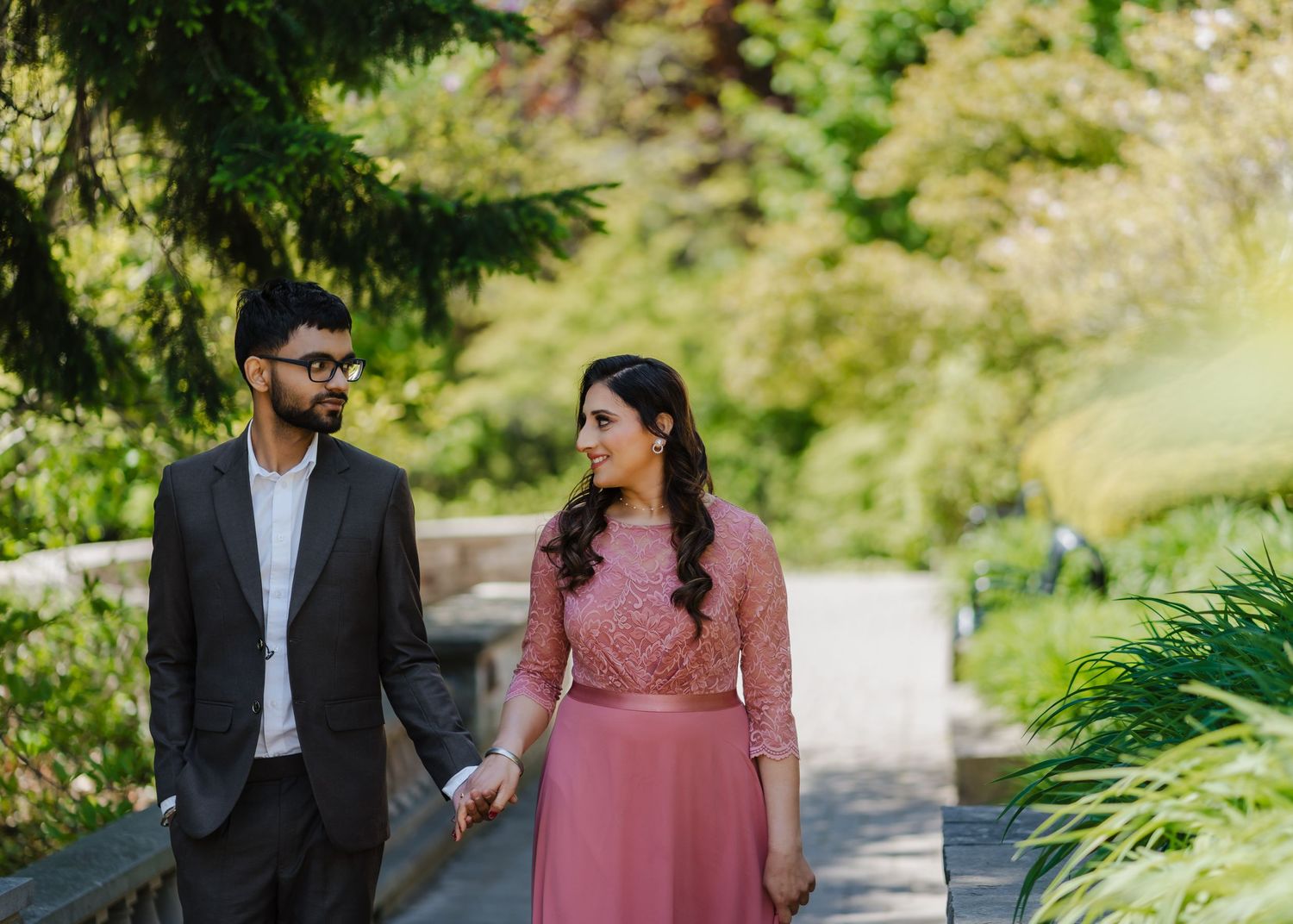 A couple walks hand in hand through a lush garden path wearing formal attire including a pink dress and dark suit.