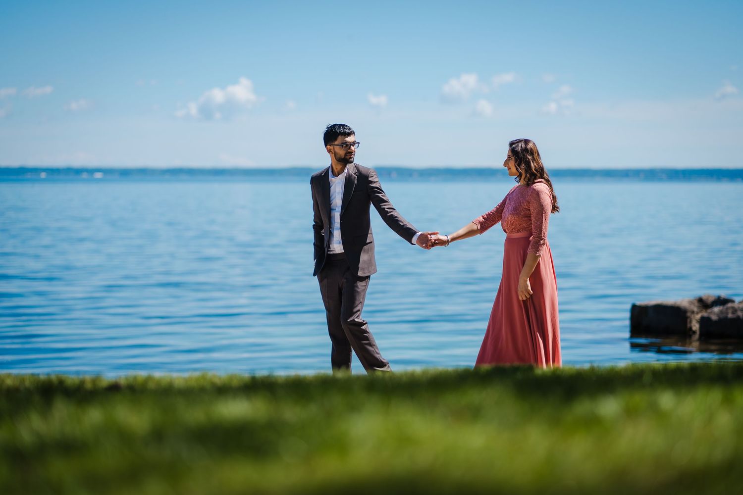 Couple in love holding hands while walking along a scenic waterfront path with blue ocean and sky in the background.