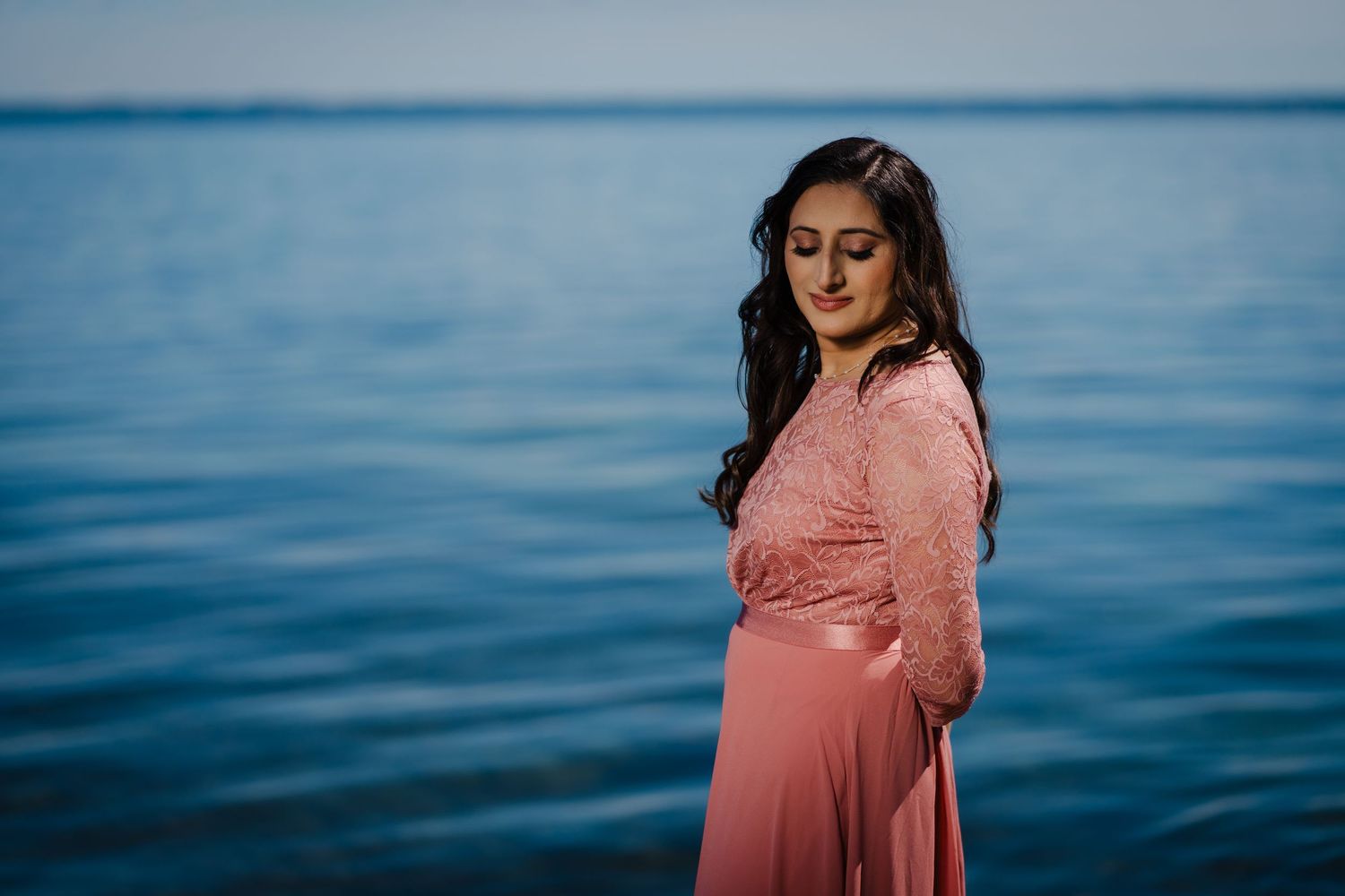 A pregnant woman in a coral dress stands by a serene blue ocean during a maternity photoshoot.