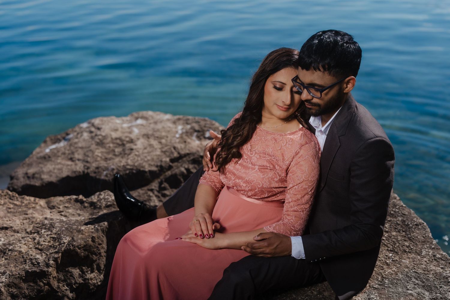 Couple in romantic embrace sits on coastal rocks with blue ocean waters in background.