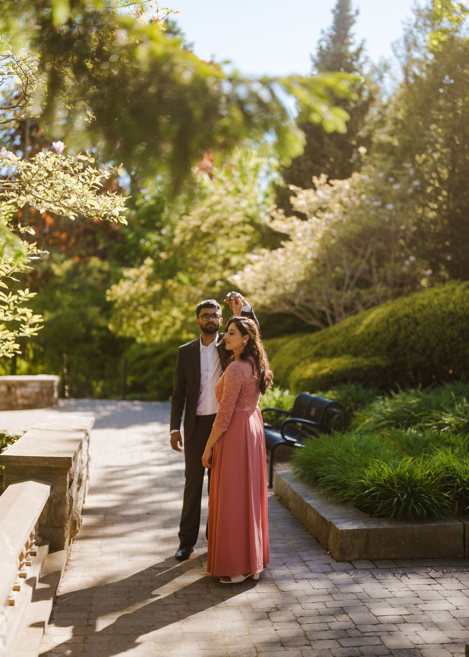 A couple in formal attire walks along a winding garden path lined with lush greenery on a sunny afternoon.