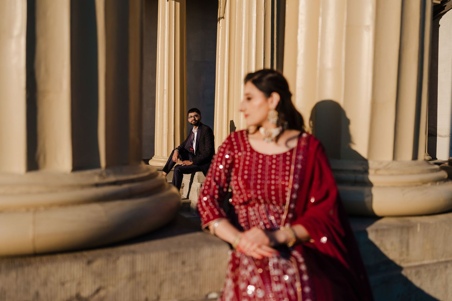 A woman in a flowing red dress poses elegantly between classical stone columns in a dramatic outdoor photoshoot.