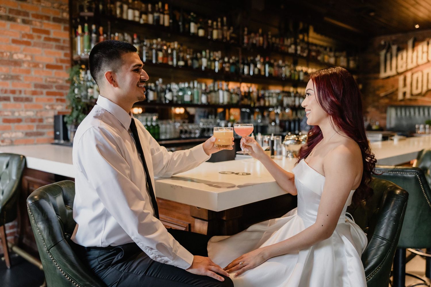 A couple in formal wedding attire shares an intimate moment at a rustic bar with exposed brick walls and wooden details.