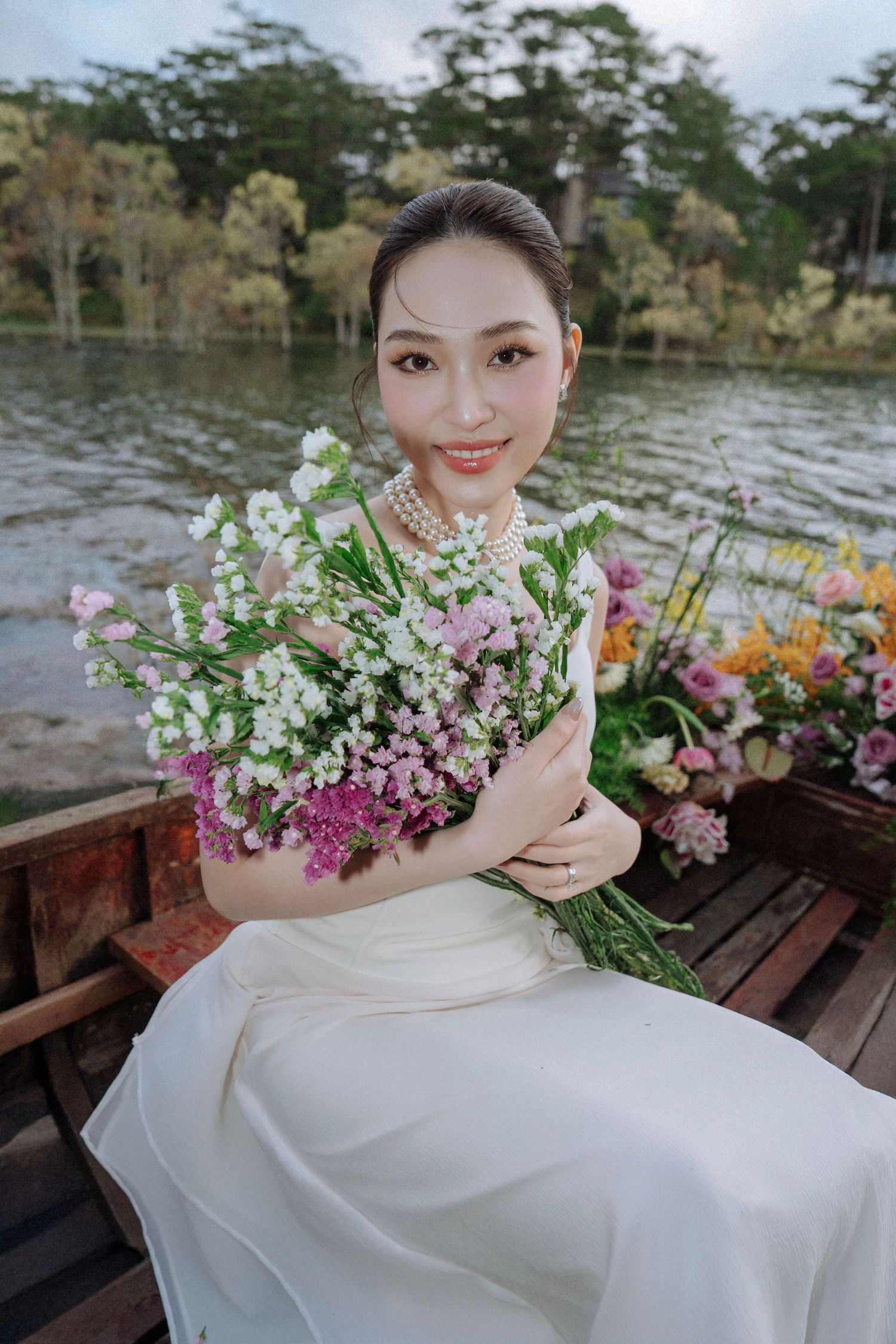 Bride in white dress holding colorful wildflower bouquet while sitting in a wooden boat on a serene lake.