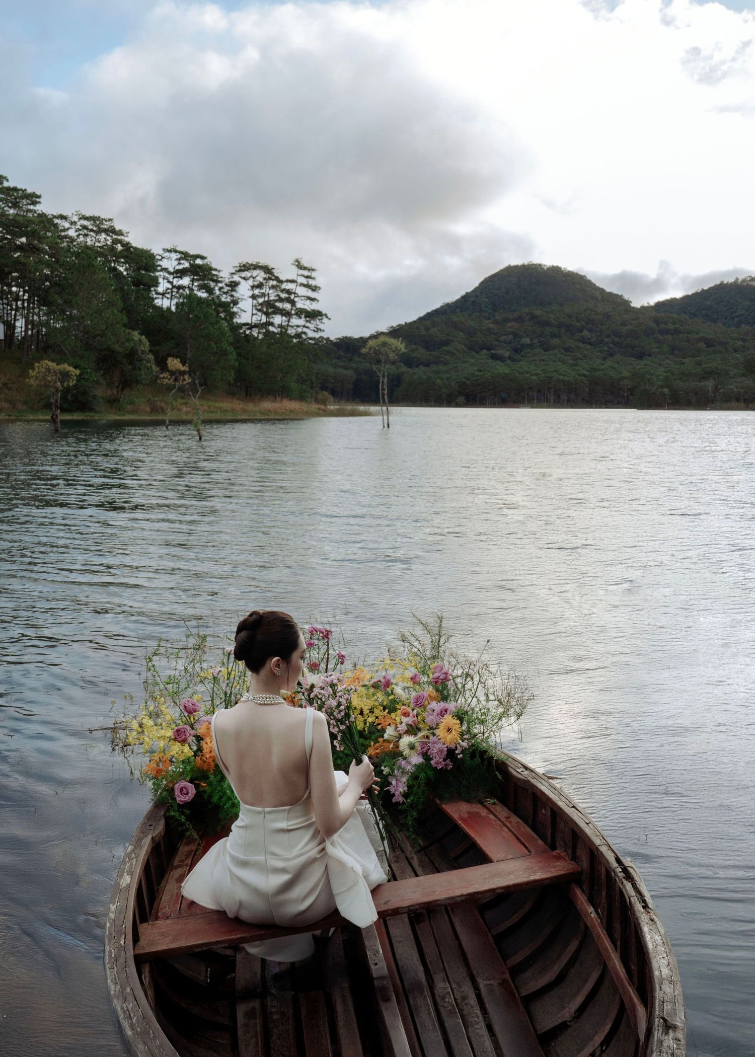 Bride in white backless dress seated in a flower-filled wooden boat on a scenic mountain lake.