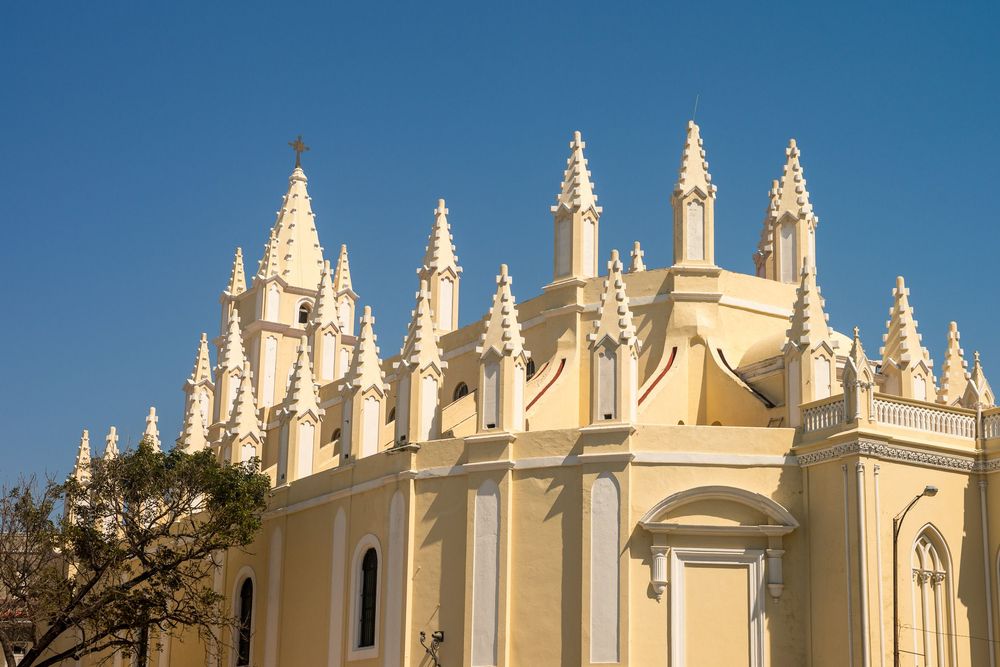 Gothic spires adorn the ornate white cathedral facade rising against a bright blue sky in tropical setting.