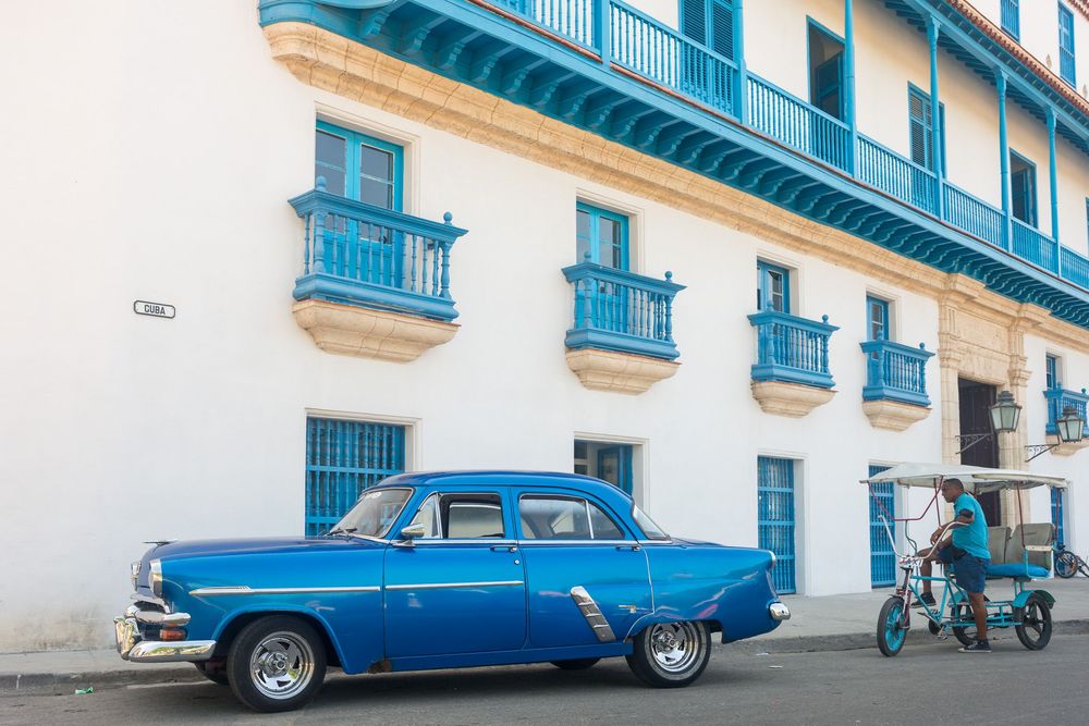 Classic blue car parked on a cobblestone street lined with white buildings and turquoise balconies in Cuba.