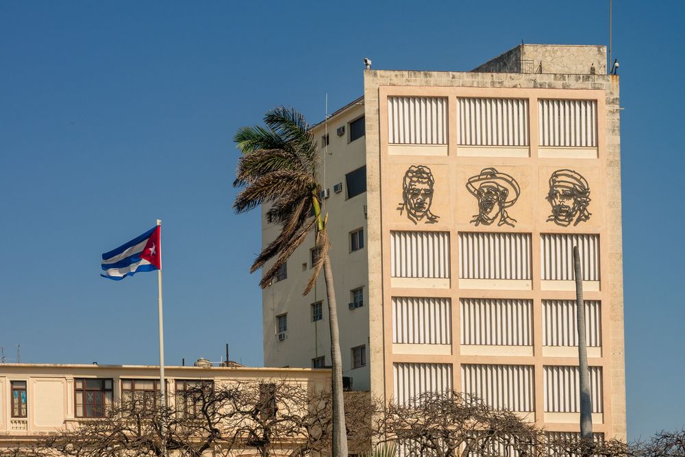 Cuban flags fly from ornate colonial buildings with balconies along a city street under bright blue sky.