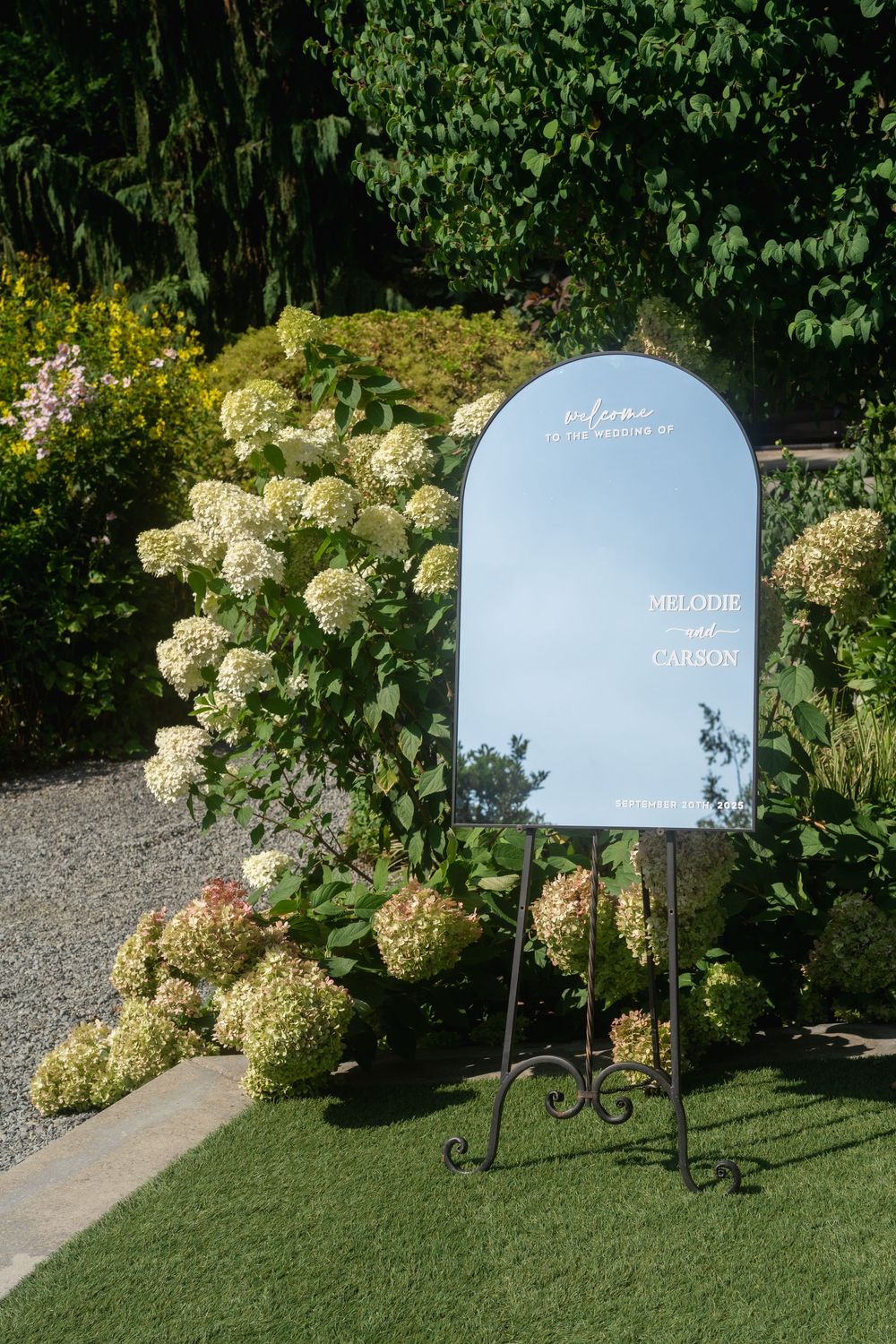 A wedding welcome sign stands elegantly on a path bordered by lush hydrangea bushes and manicured lawn.