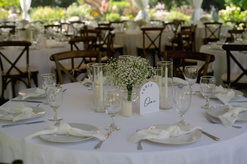 Elegant wedding reception tables set with white linens, floral centerpieces and rustic wooden cross-back chairs.