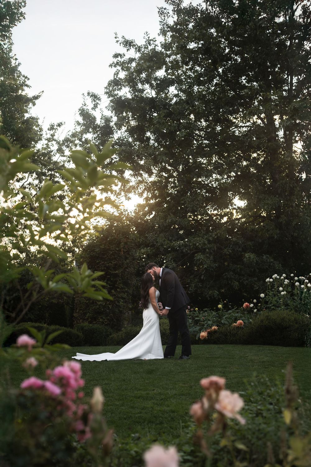 Newlyweds share intimate moments against a lush garden backdrop with pink flowers and setting sun.