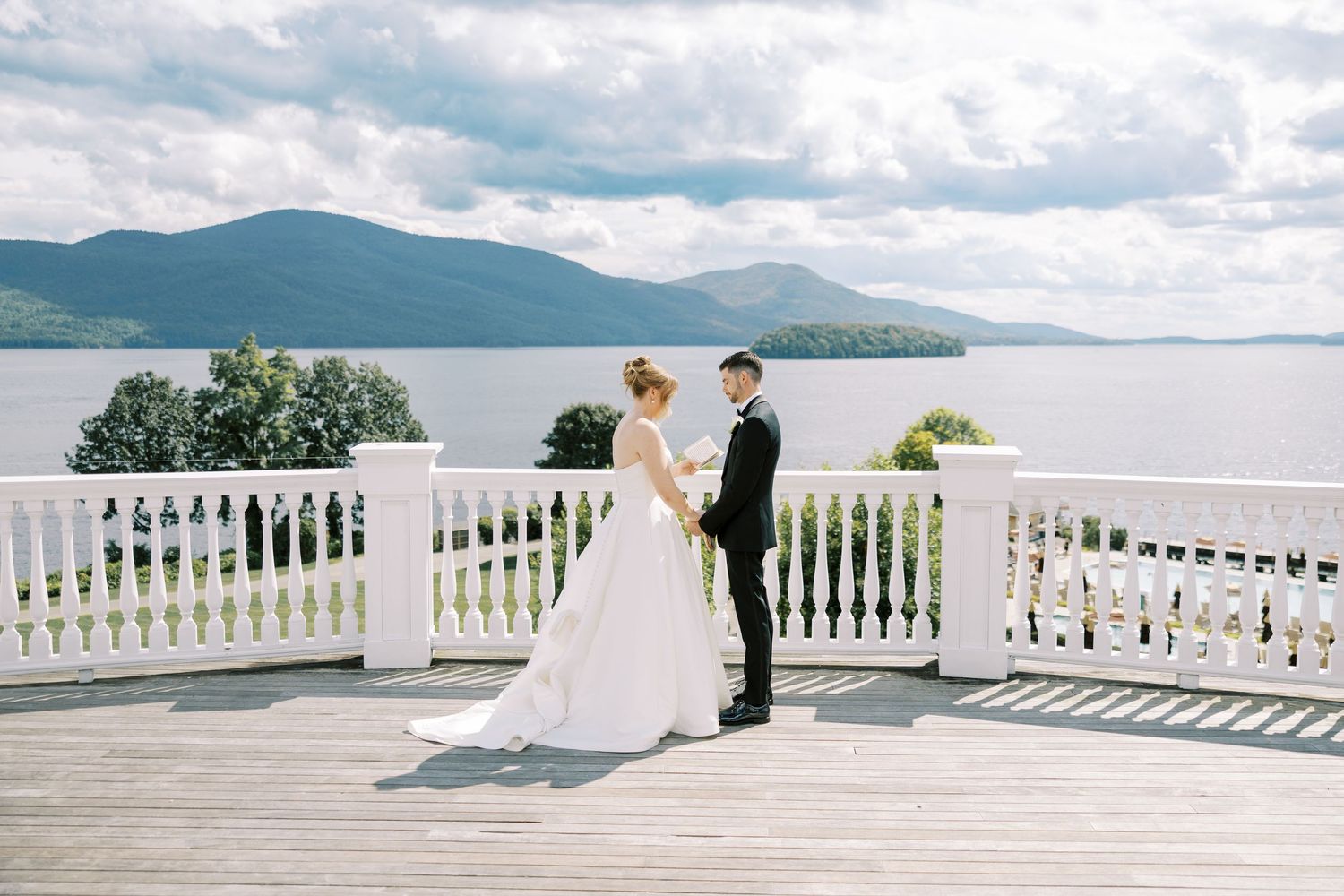 A couple in wedding attire stands on a white balcony overlooking a serene lake with mountains in the distance.