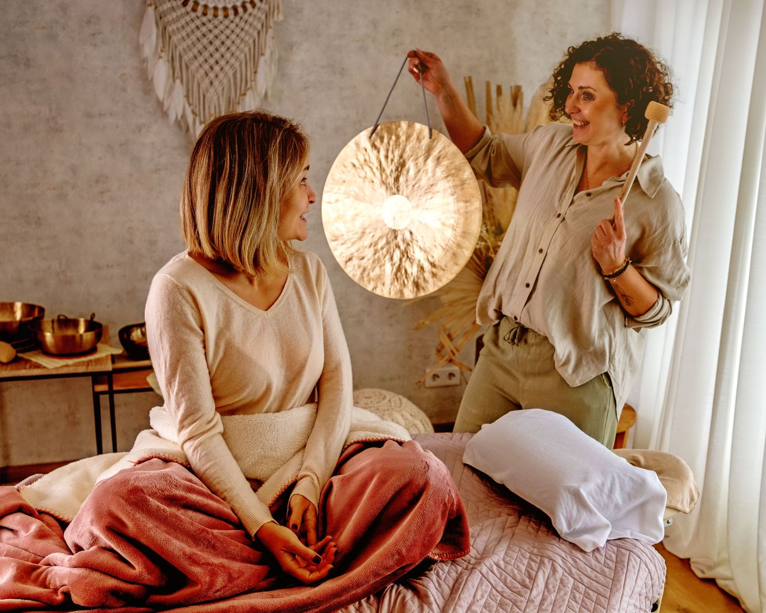 Two women in a cozy boho bedroom, one holding a glowing lamp during a relaxing sound healing session.