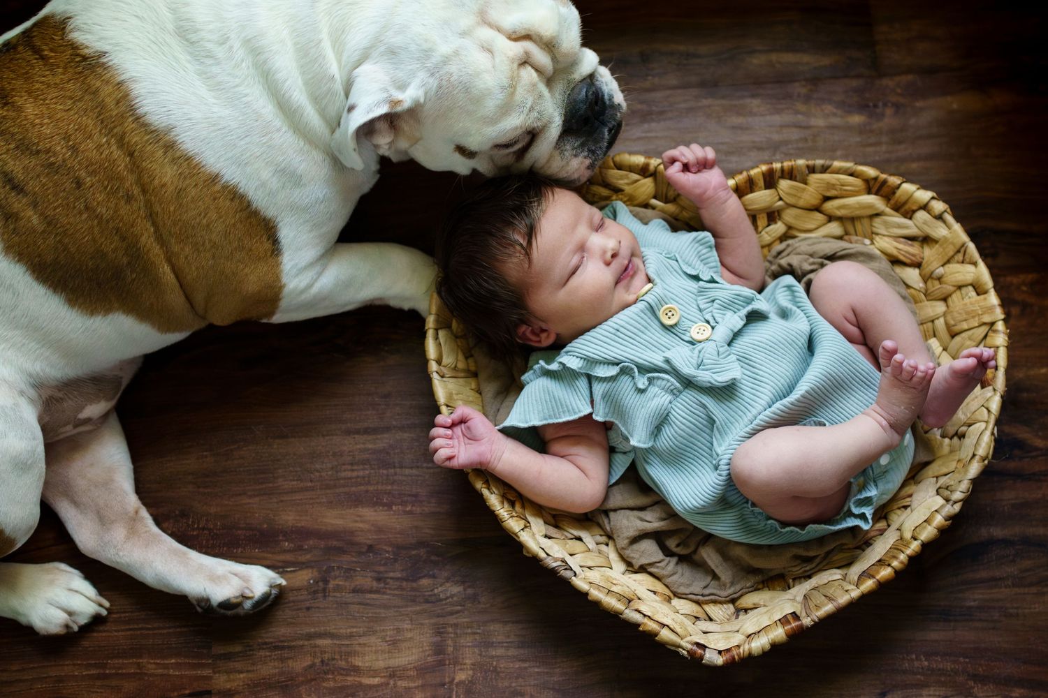 Peaceful sleeping baby in a woven basket on wooden floor wearing light blue clothing posed during in-home newborn session.
