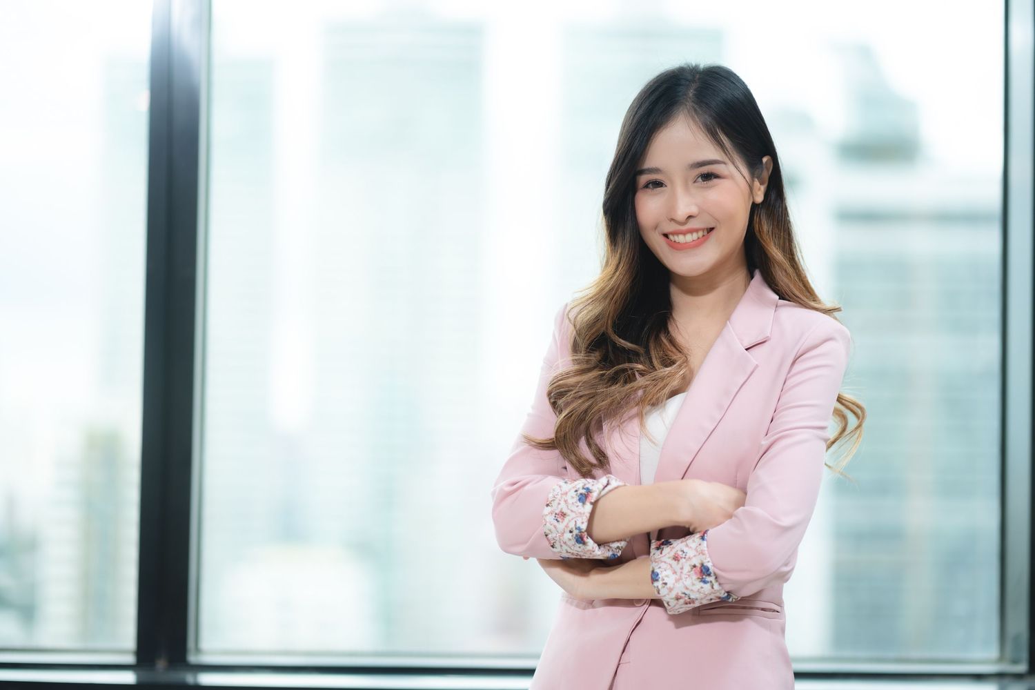 Professional wearing a pink blazer stands confidently in a modern office setting with large windows.