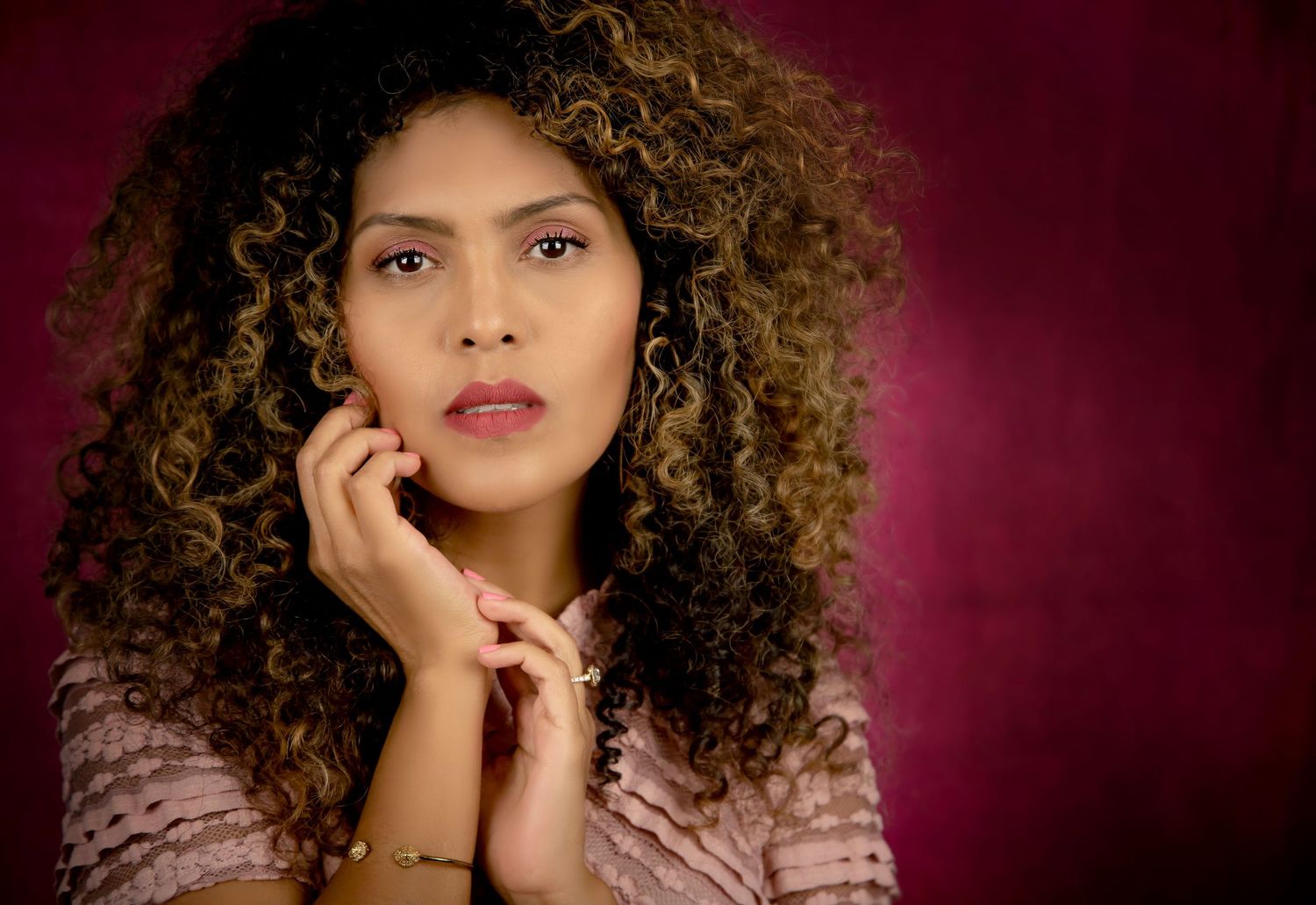 Elegant portrait with curly hair against burgundy backdrop showing contemplative expression.