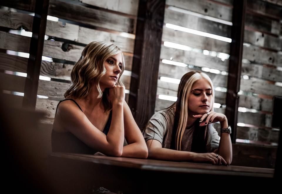 Two women in pensive poses lean on wooden table in dimly lit room with slatted wall background.