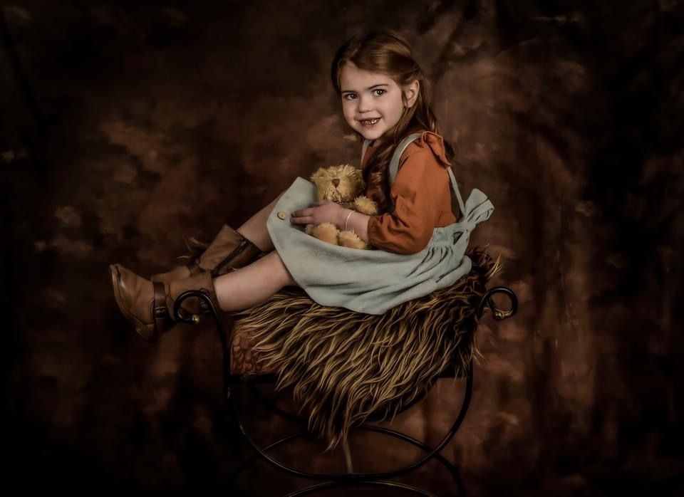 Fine art portrait in dark browns showing a seated figure holding a teddy bear on a rustic straw chair.