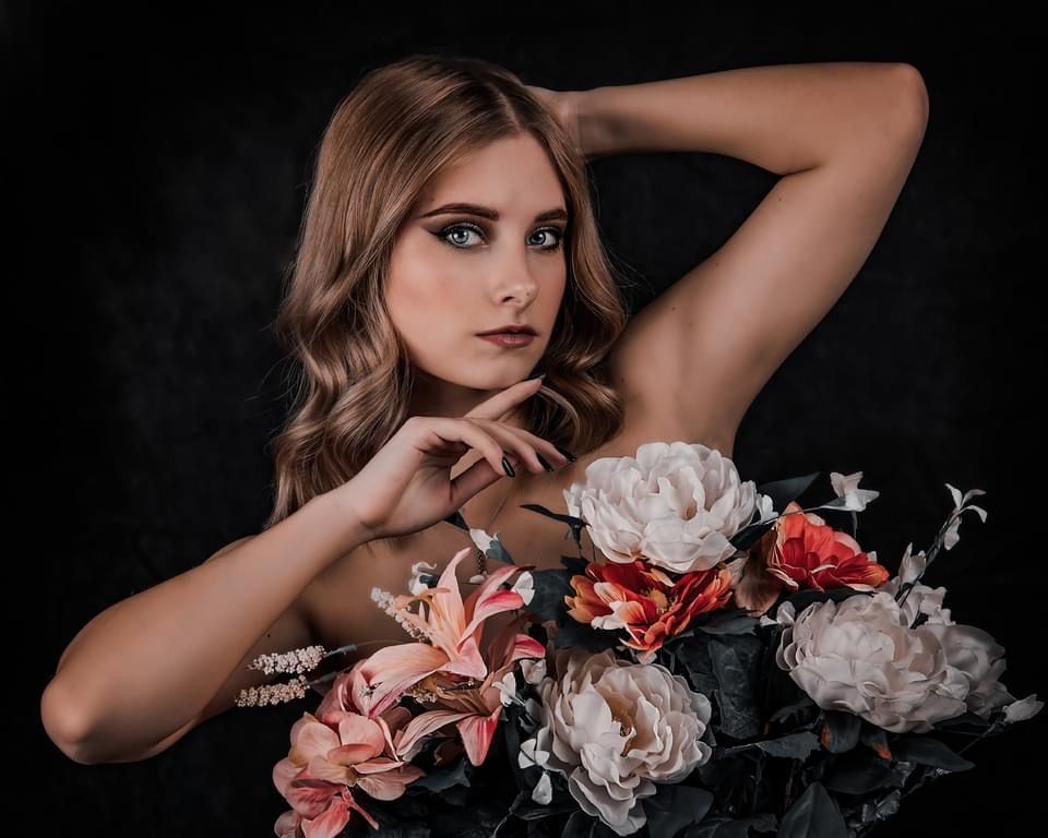 Dramatic beauty portrait with pink and white flowers against dark background showing elegant makeup and styling.
