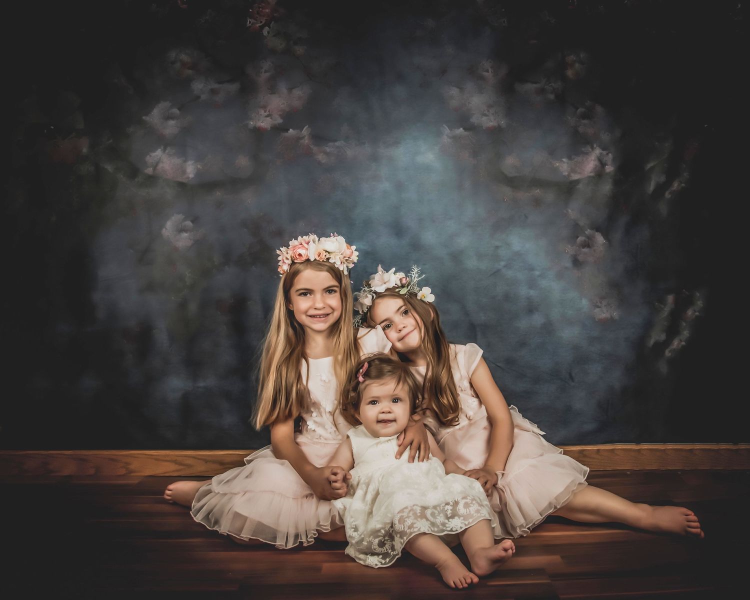 Three children in white dresses pose together on wooden floor during elegant studio photoshoot.