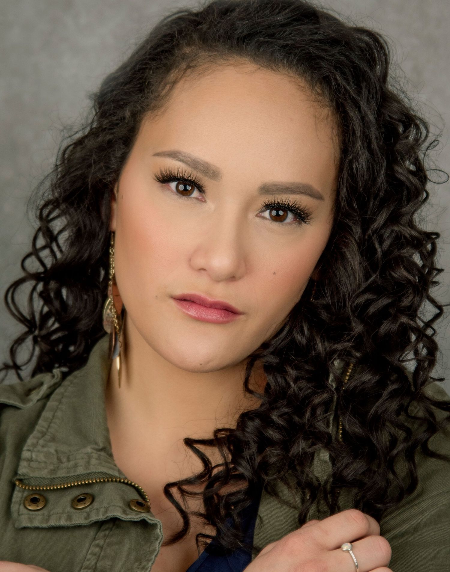 Professional portrait shot with dramatic lighting showing curly dark hair, wearing an olive green jacket and dangling earrings.