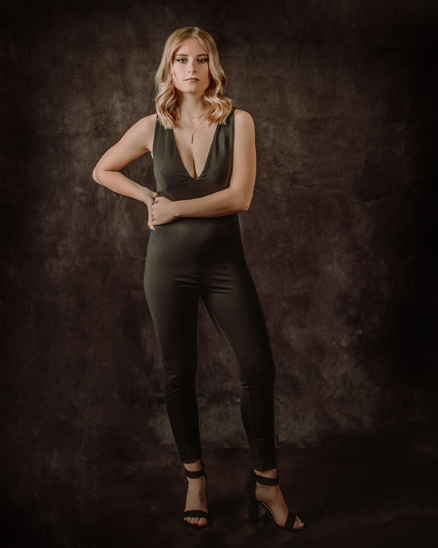 Fashion portrait in moody dark studio setting shows model in black jumpsuit striking confident pose against textured backdrop.