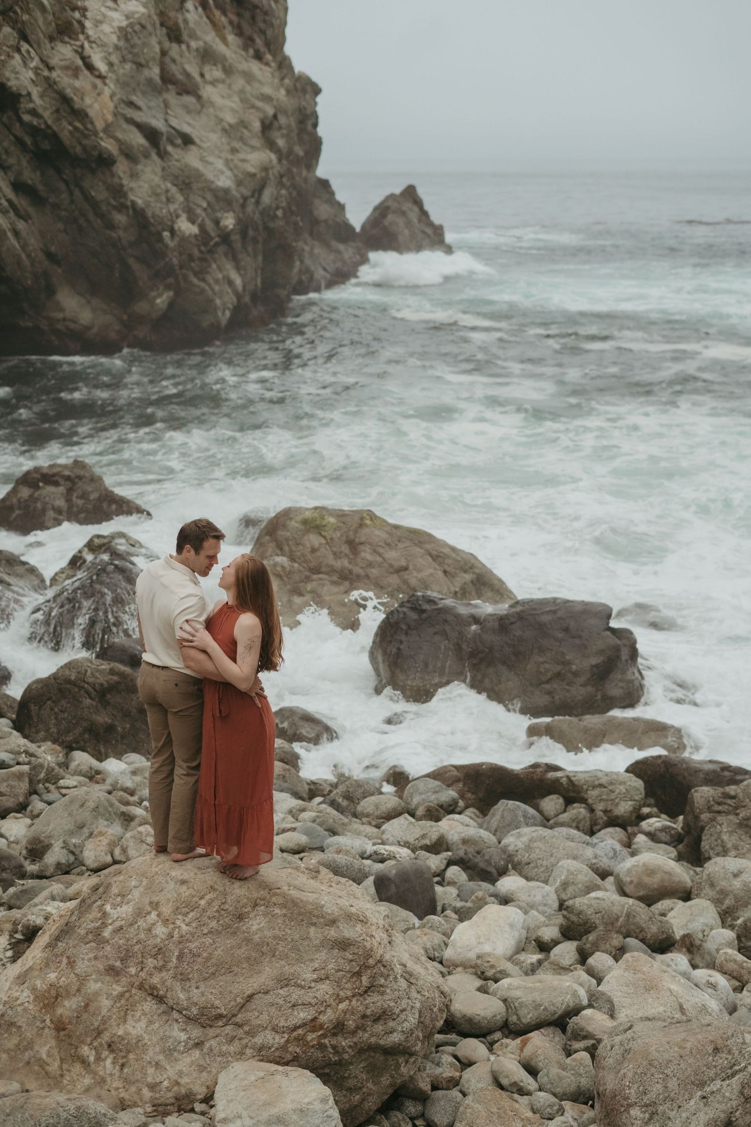 Two people sit together on coastal rocks as waves crash against the rugged shoreline in moody overcast weather.