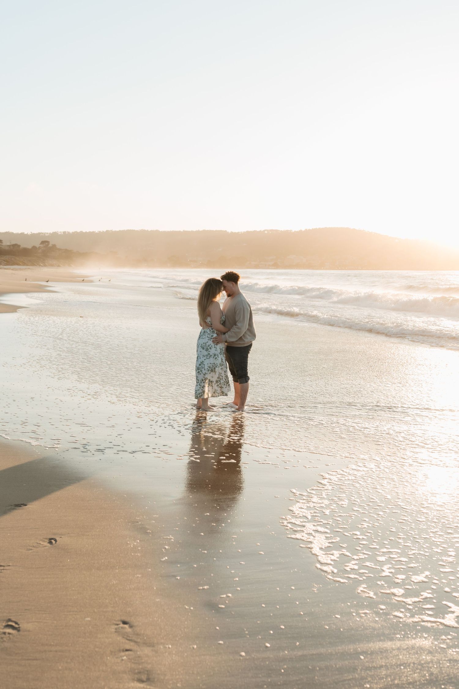 A couple embraces on a golden beach at sunset with waves gently washing over their feet.